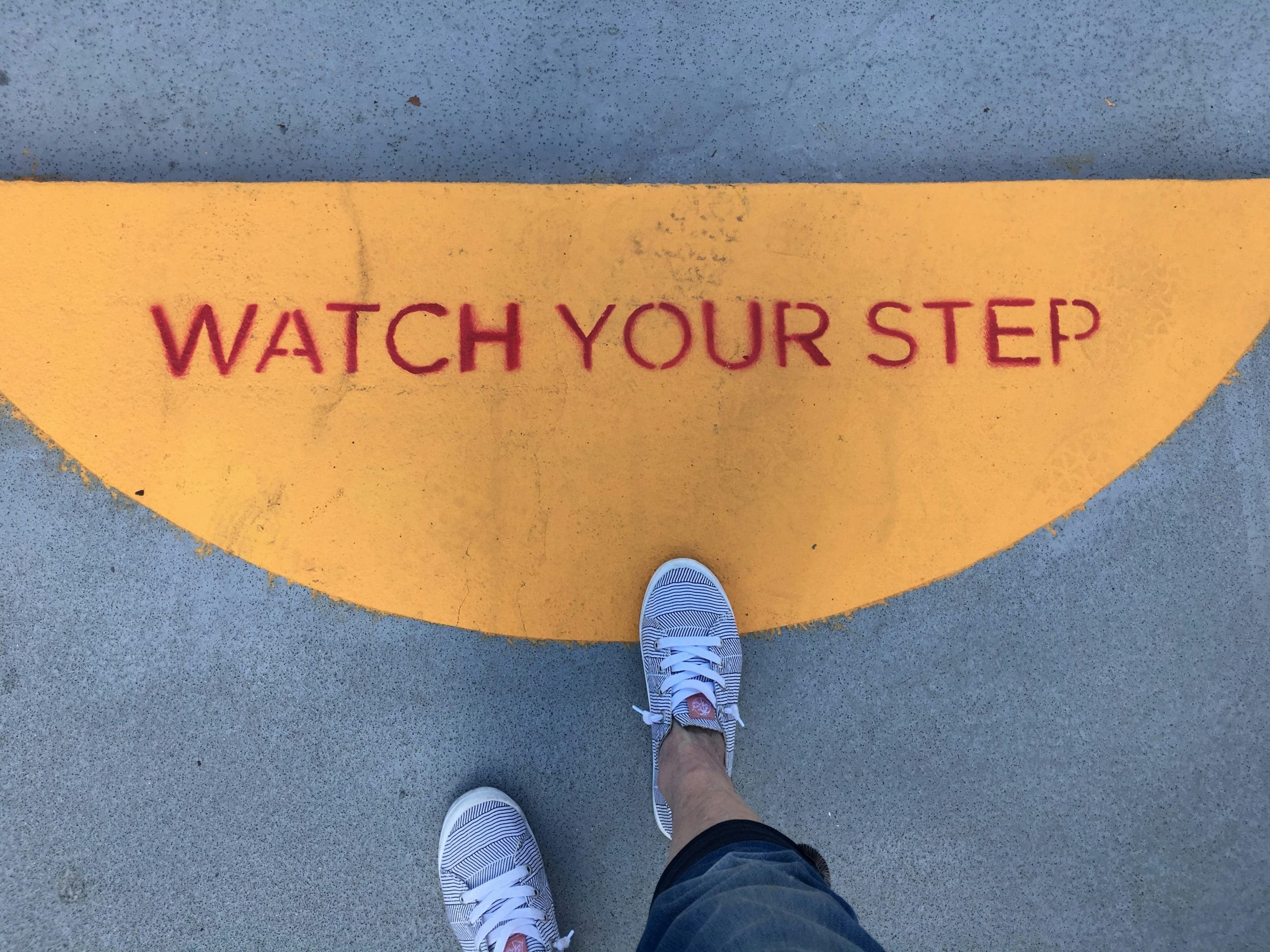 Ступни в кроссовках на бетоне рядом с жёлтой линией с надписью "WATCH YOUR STEP".
